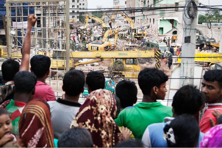 Elf Jahre nach Rana Plaza: Textilfabriken sicherer, Auslöser der Katastrophe bleiben 6 Eine Gruppe Menschen - von hinten zu sehen - stehen vor einem Gitter. Dahinter sind die Trümmer eines eingestürzten Gebäudes zu sehen.