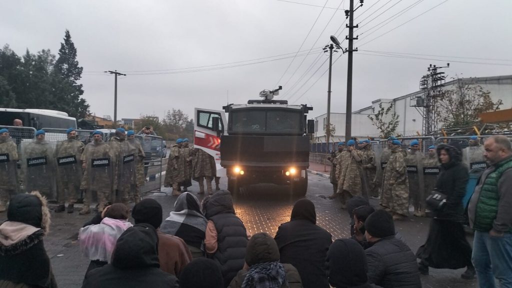 Soldaten und ein Militär-Lkw stehen einer Gruppe Menschen gegenüber. Der Himmel ist grau.