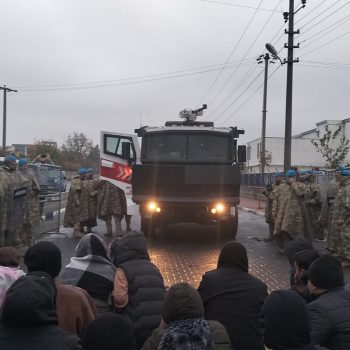 Soldaten und ein Militär-Lkw stehen einer Gruppe Menschen gegenüber. Der Himmel ist grau.