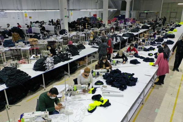 An langen Tischen sitzen Menschen nebeneinander und nähen. Sie tragen Masken.