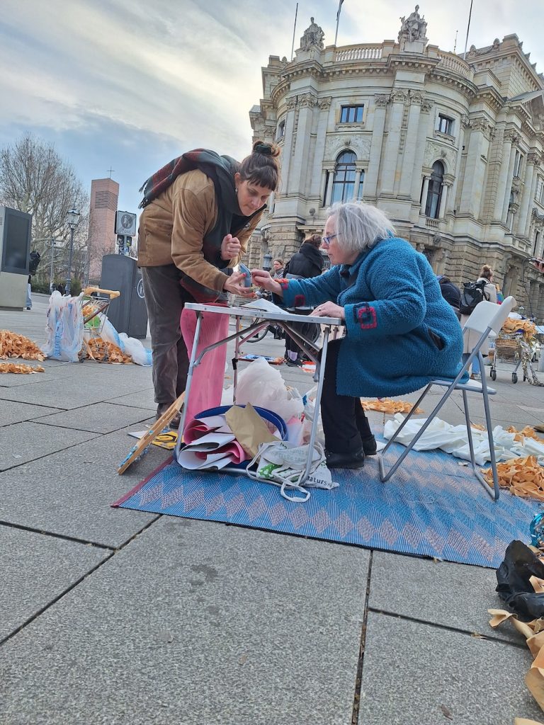 "Ein Wochenende voller Erkenntnisse, Gemeinschaft und Aktivismus" - Lena berichtet über ihr erstes Aktionstreffen 3 Eine Frau sitzt auf einem Platz an einem kleinen Tisch. Eine andere beugt sich über den Tisch.