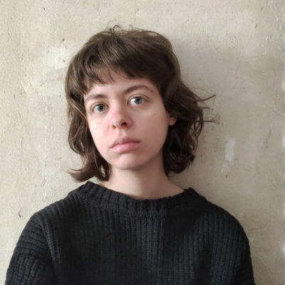Eine junge Frau in schwarzem Pulli steht vor einer grauen Wand.